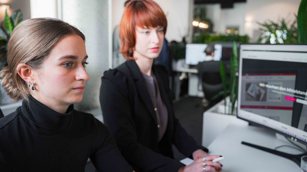 Zwei Frauen sitzen an einem Schreibtisch und betrachten gemeinsam einen Computerbildschirm.